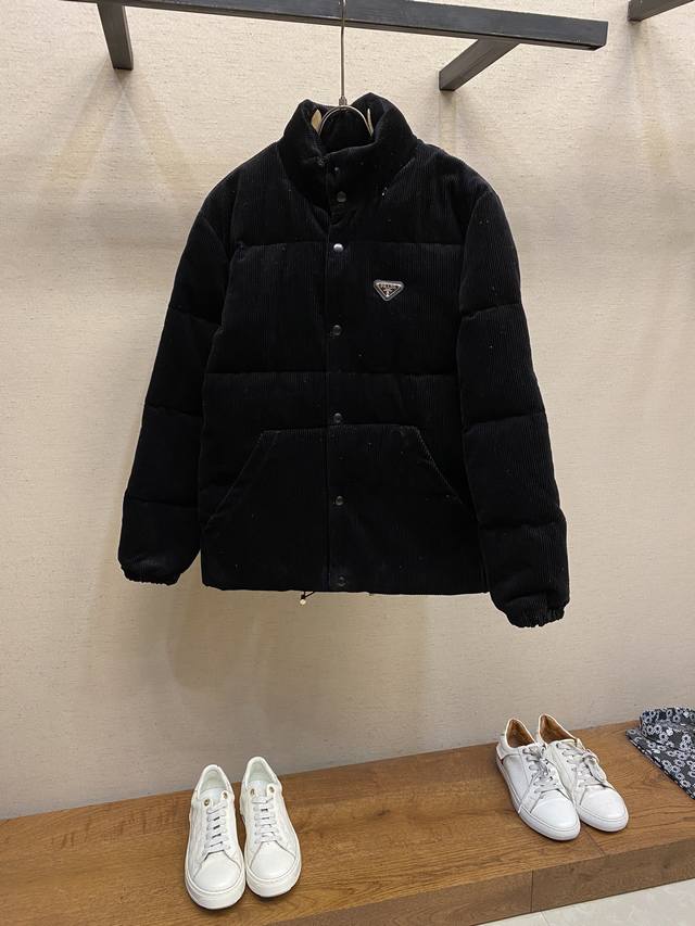 Prada 24Fw 灯芯绒立领羽绒服 这款短裁灯芯绒羽绒 蕴藉prada的混搭个性 金属三角形徽标展现标志性元素 灯芯绒为生产痛点 所产生的 面料光差 顺毛差